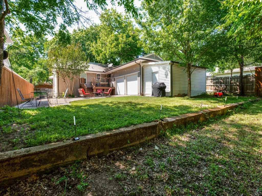 Backyard with green lawn, trees, a patio area with chairs, and a small building with a white garage door.