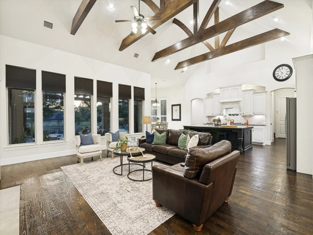Living room with dark wood floor, white walls, large windows, and a vaulted ceiling with exposed wooden beams. Features include a brown leather sofa, white armchairs, a patterned area rug, and a black kitchen island with white cabinetry in the backgr
