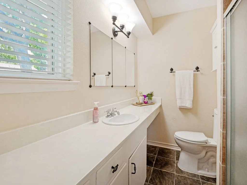 A bathroom with a white vanity, a sink, and a mirror, a window with blinds, a toilet, a towel rack with a towel, and a shower stall, decorated with small plants and toiletries.