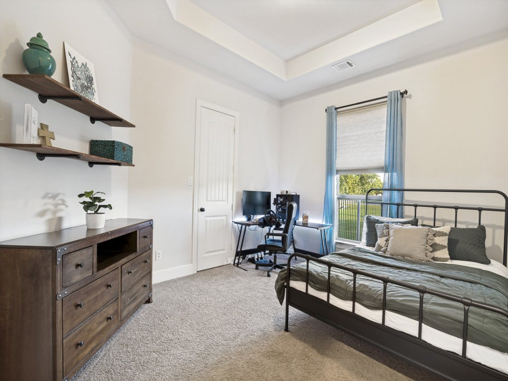 A tidy bedroom with a metal bed frame, a window with blue curtains, a desk with a computer, and a wooden dresser with shelves above it decorated with vases, artwork, a cross, and a box.