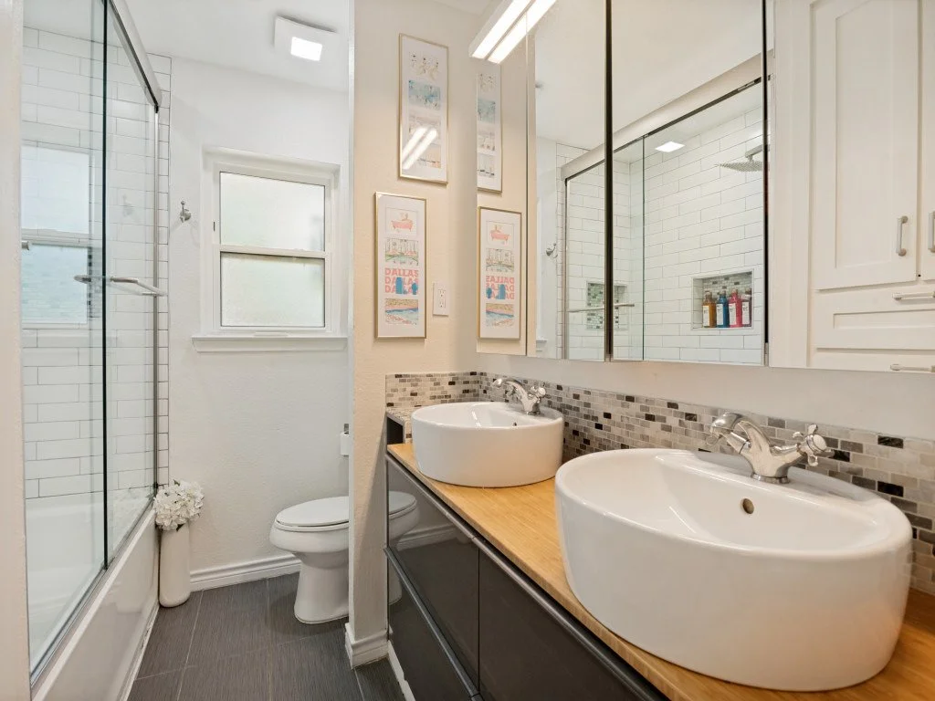 A modern bathroom with double vessel sinks on a wooden vanity, a large mirror above, a shower with glass doors, white subway tile walls, a small window, and framed artwork on the wall.