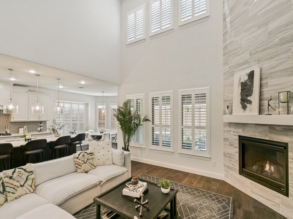 Living room with a white sectional sofa, coffee table, fireplace, and wooden floors, with large windows with white shutters and modern decor.