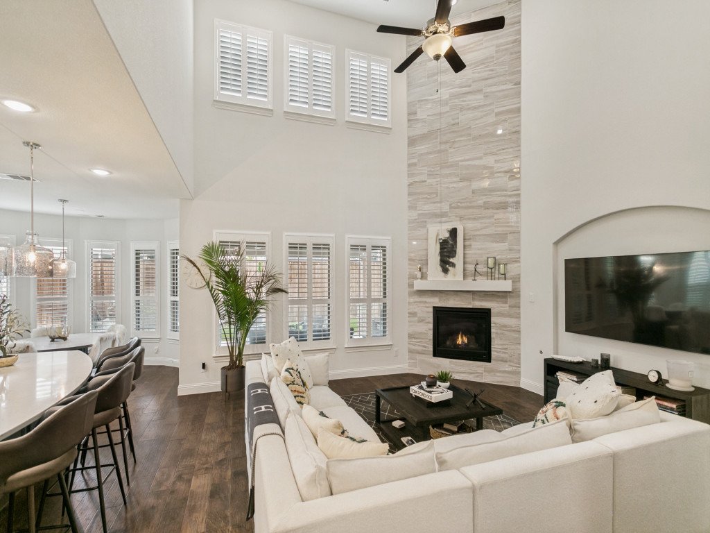 A spacious living room with a high ceiling featuring a ceiling fan, a fireplace with a textured wall, a large flat-screen TV, and a white sectional sofa. There are multiple windows with white shutters, and a dining area with a curved table and chairs visible in the background.