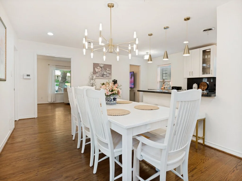 Dining room with a white table, six white chairs, hanging ceiling lights, wooden flooring, and a view into the kitchen and living area with a TV and window.