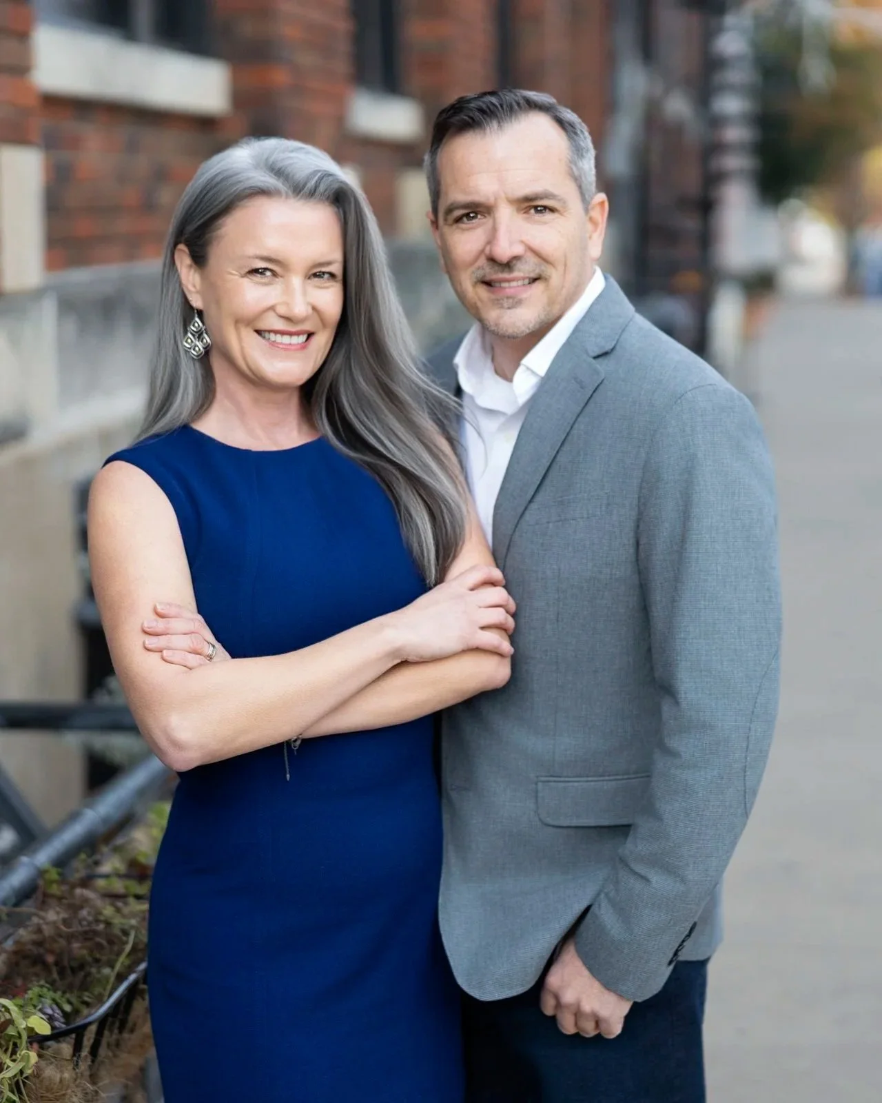 A smiling middle-aged woman with long gray hair wearing a sleeveless navy blue dress, standing outdoors with her arm crossed. Beside her, a smiling middle-aged man with short dark hair and a hint of gray, wearing a light gray suit jacket over a white shirt, stands on a city street with brick buildings in the background.