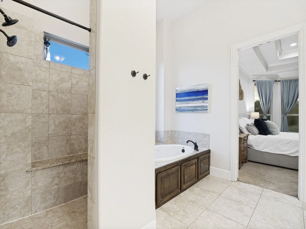Bathroom with a walk-in shower, a bathtub beneath a small window, and an adjacent bedroom with a bed and blue curtains.