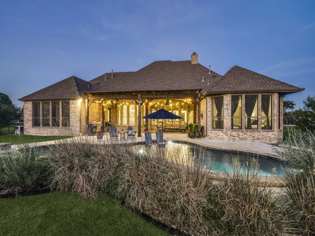 A backyard view of a brick house with large windows, a covered patio, outdoor furniture, string lights, a swimming pool, and dry plants in the foreground during evening with a blue sky.