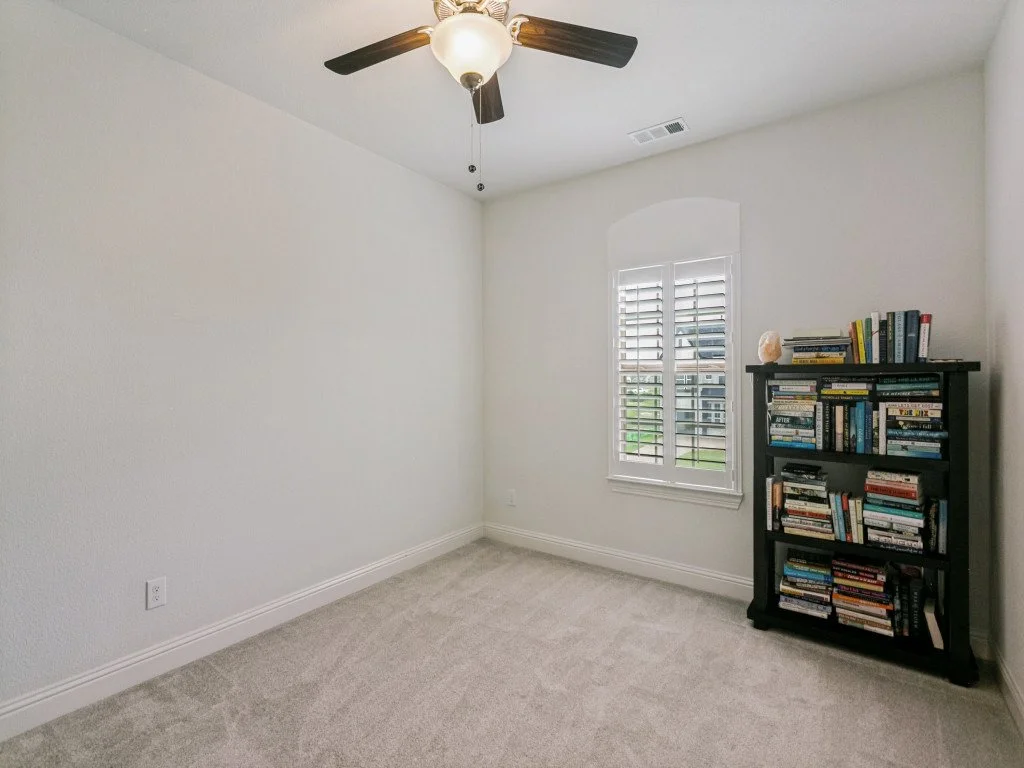 Empty room with a ceiling fan, a window with shutters, and a black bookshelf filled with books.