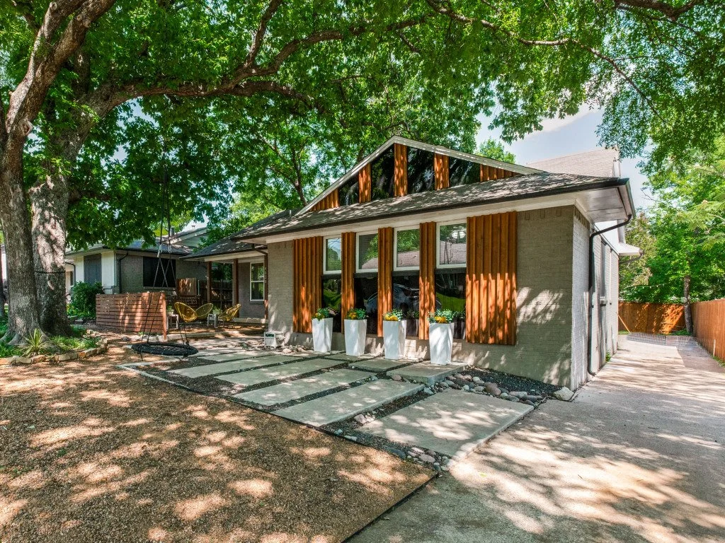 The backyard of a house with a large tree providing shade. The house has a modern design with vertical wooden slats on the exterior and large windows. There is a stone pathway leading to the house, potted plants, a swing, and outdoor seating, creatin