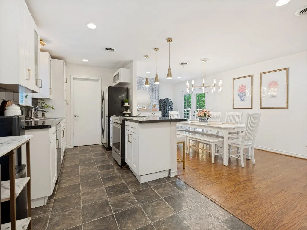 Bright, modern kitchen and dining area with white cabinetry, black countertops, stainless steel appliances, and a white dining table with chairs. Gold and pendant lighting, framed floral artwork, and wood and tile flooring.