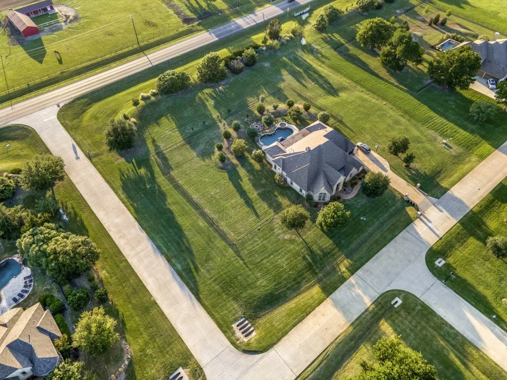 Aerial view of a suburban neighborhood showing a house with a backyard pool, surrounded by greenery and paved walkways, with adjacent houses, roads, and lawns.