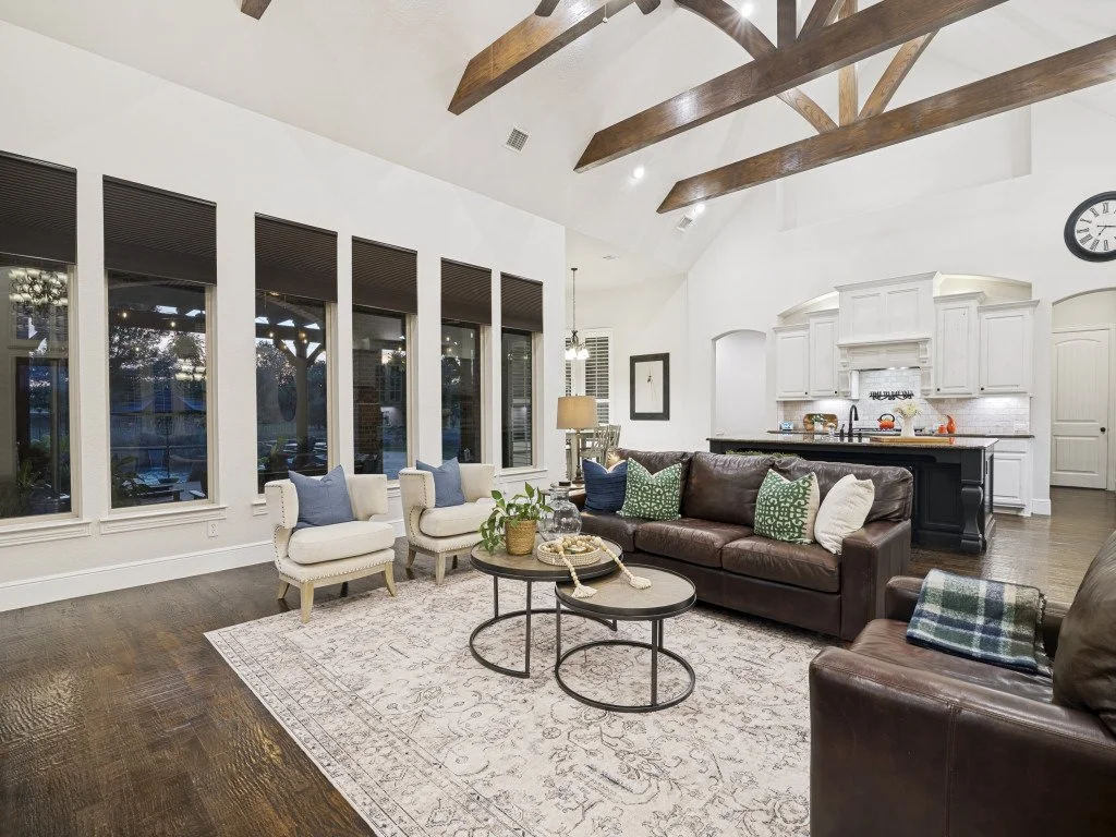 Living room with high vaulted ceiling, wooden beams, white walls, large windows with blinds, and a kitchen in the background. It has a brown leather sofa, white armchairs, a patterned area rug, nested coffee tables, and decorative pillows, with a clo