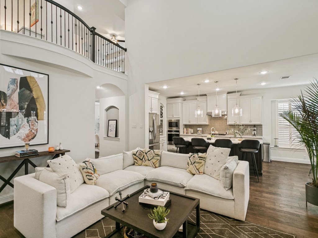 Open-concept living room and kitchen in a modern home with white walls, a sectional white sofa, and a wooden coffee table, with the kitchen featuring white cabinetry, stainless steel appliances, a gray backsplash, and a breakfast bar with black stool