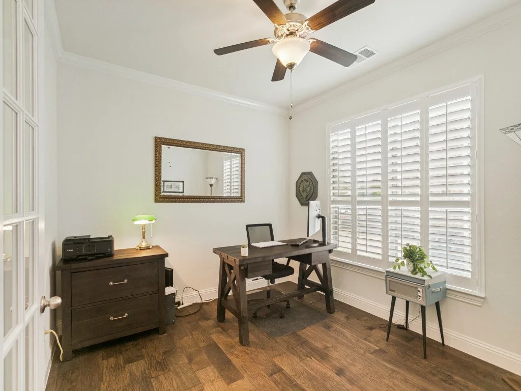 A small home office with a desk, a black office chair, a dresser with a green banker lamp, a wall mirror, and a window with white shutters. Ceiling fan hanging from the ceiling, wooden floor, and a potted plant on a table.