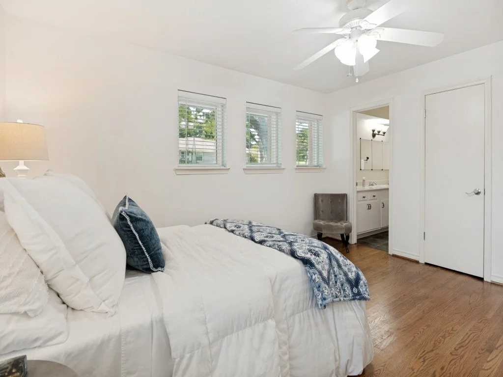 A bedroom with white walls, a white bed with pillows, and a navy blue throw blanket. There are three windows with blinds, a beige lamp, a gray upholstered chair, and a small bathroom with a sink and mirror visible through an open door. A white ceilin
