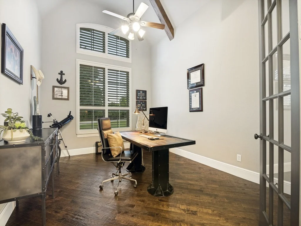 Home office with a large wooden desk, black office chair, desktop computer, window with plantation shutters, ceiling fan, framed artwork and certificates on the wall, telescope, and decorative items on a side table.