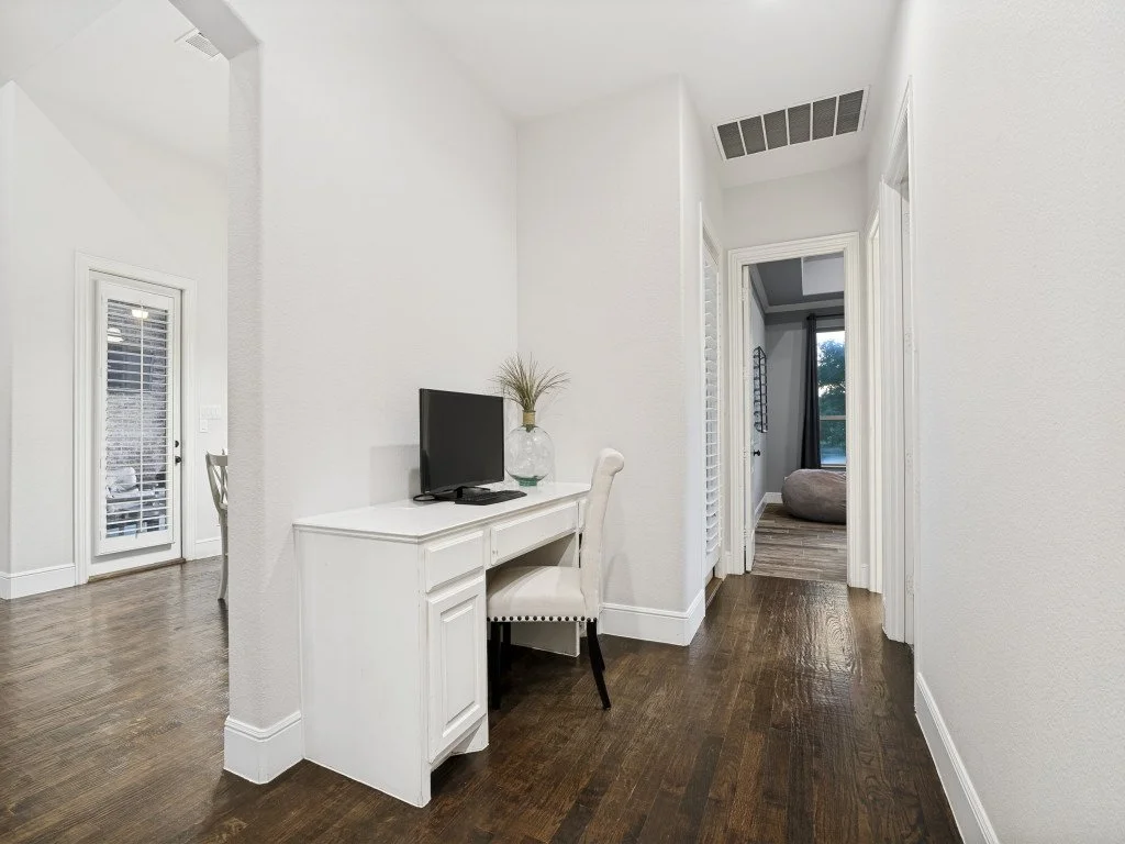 A hallway with white walls and dark hardwood flooring, featuring a computer desk with a monitor and vase with plant, and doorways leading to other rooms with visible furnishings.