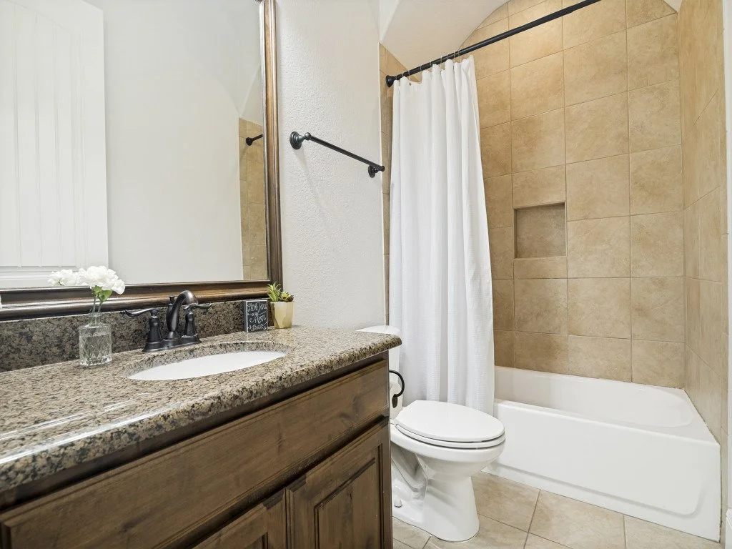 Bathroom with a granite countertop, a mirror, a small potted plant, a vase with flowers, a black faucet, a toilet, a bathtub with beige tile walls, a shower curtain, and a small built-in niche in the shower wall.