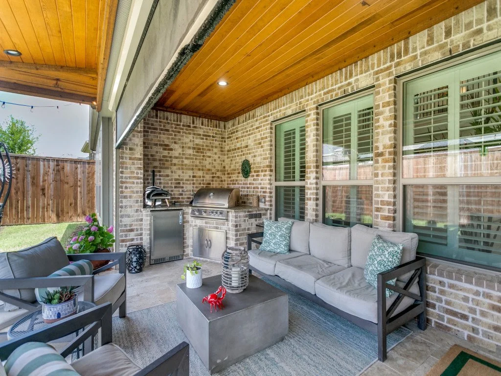 Covered outdoor patio with brick wall and wooden ceiling, outdoor grill, sofa, armchair, outdoor rug, and decorative items.