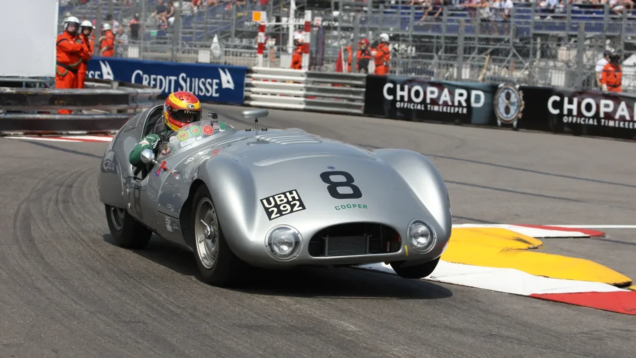 A vintage silver race car with the number 8 on the front speeding on a racetrack with safety barriers and spectators in the background. The driver is wearing a red helmet. Chris Ward Monaco Historic 2024. Legacy Outlaws Racing Team.