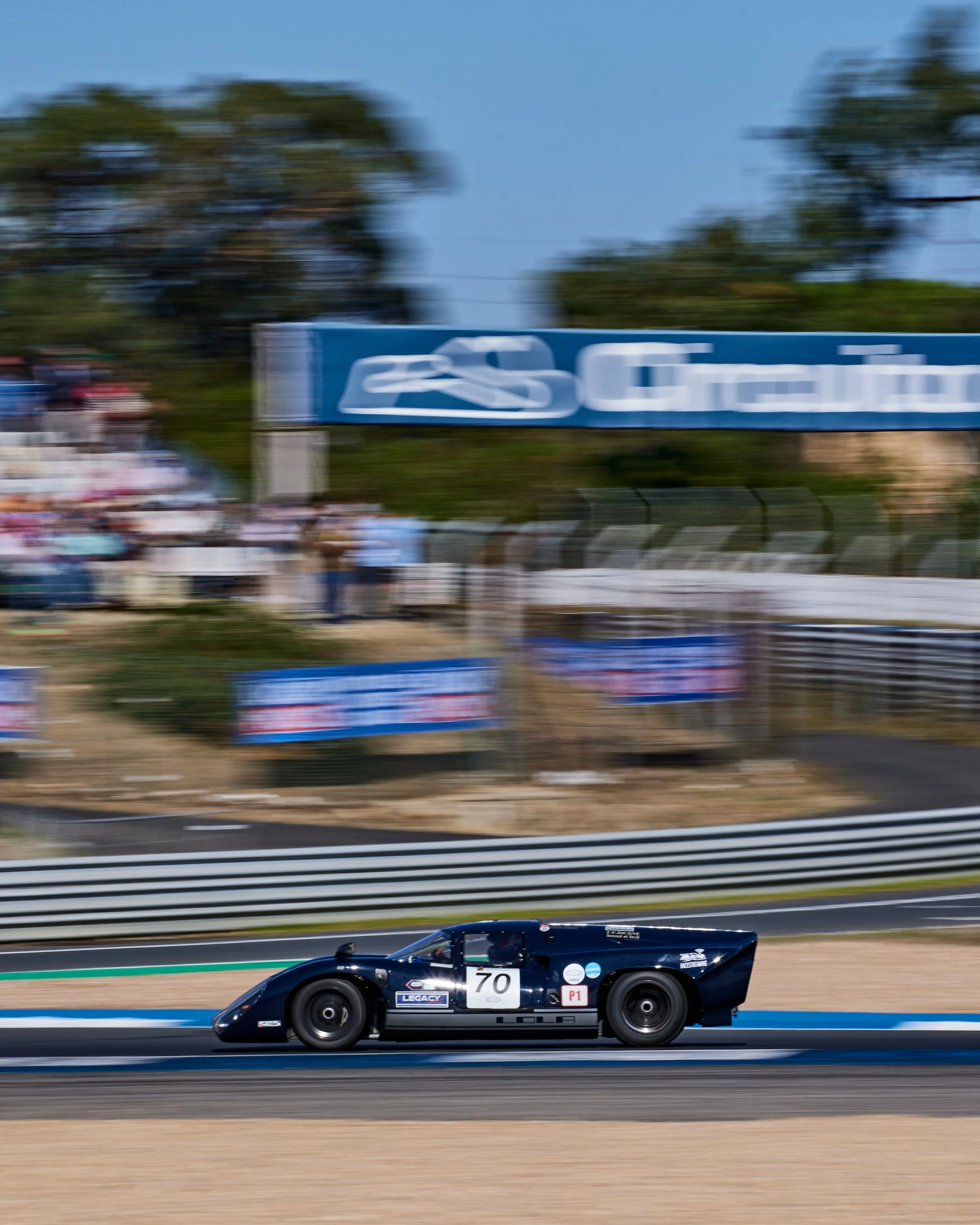 A black race car with the number 70 racing on a track, with a blurred background of trees, billboards, and people watching the race. Legacy Outlaws Racing Team. Peter Auto Estoril Classic 2025. Chris Ward and Jan Magnussen. 