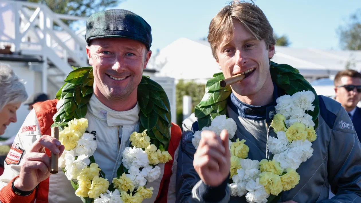 Two race car drivers celebrating with leis and cigars, one with a cigar in his mouth, outdoors with a crowd and white tents in the background. Chris Ward and Gordon Sheddon. Goodwood Revival. Legacy Outlaws Racing Team.