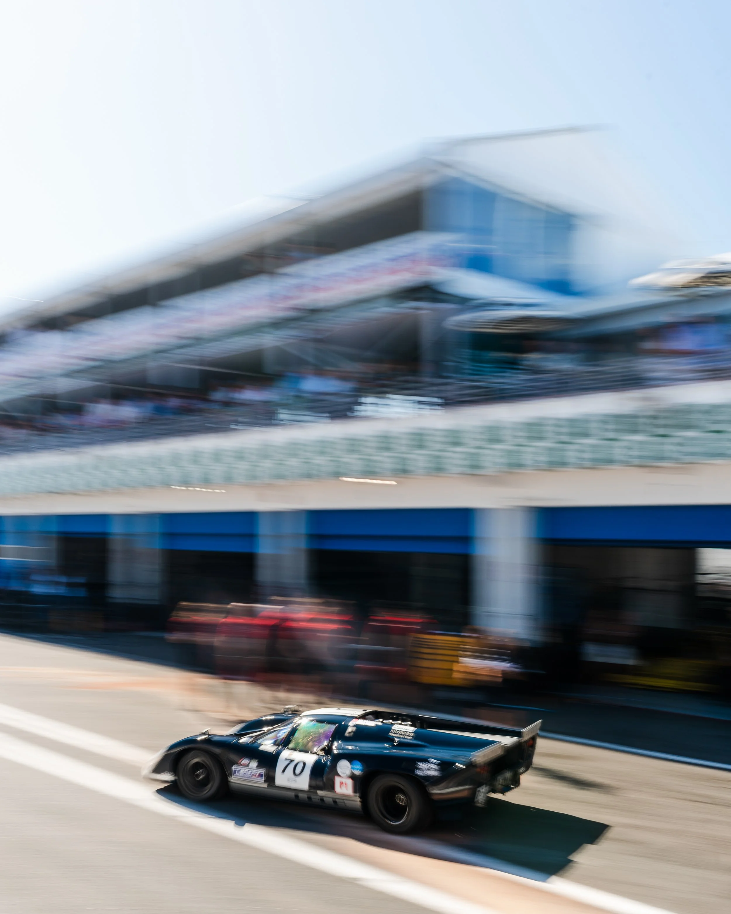 A black race car numbered 70 speeding on a race track, with blurred grandstands and structures in the background. Lola T70 MkIIIB Peter Auto Estoril Classic 2025. Legacy Outlaws Racing Team.