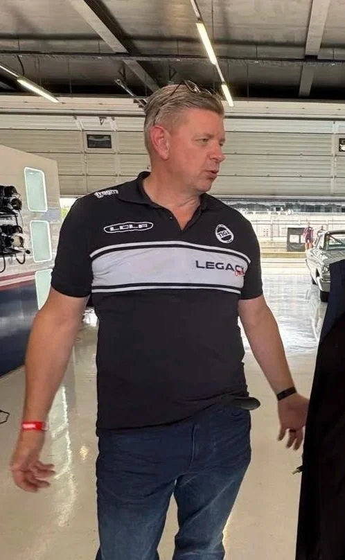 A man in a black and white racing shirt standing inside a garage or pit area, possibly at a racetrack. Lola T70 MkIIIB Motor Racing Legends Masters Silverstone Classic 2025. Legacy Outlaws Racing Team.