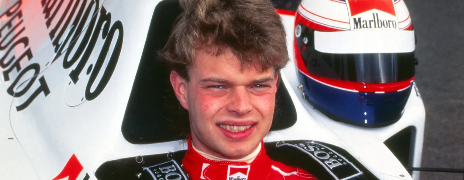 Close-up of a man with styled brown hair smiling, sitting in a race car with a white helmet nearby, on a racing track. Jan Magnussen. Legacy Outlaws Racing Team.