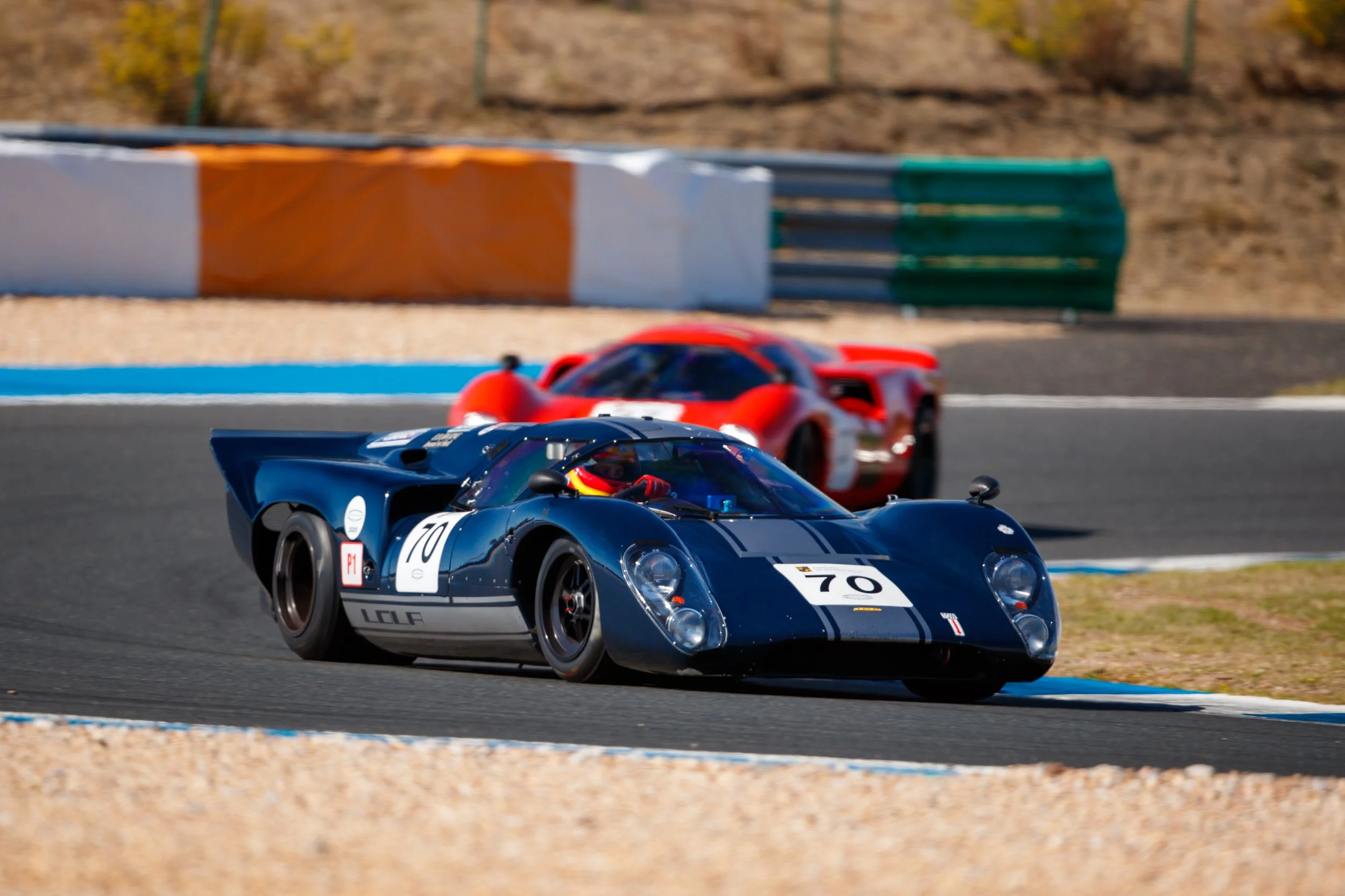 A black vintage racing car with the number 70 on it, leading a red race car on a racetrack. The scene takes place on a sunny day with barriers and gravel along the track. Legacy Outlaws Racing Team. Peter Auto Estoril Classic 2025. Chris Ward and Jan