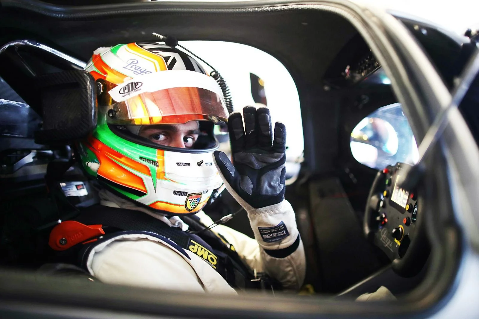 A race car driver sitting in a race car cockpit, wearing a colorful helmet and racing gloves, waving to the camera. Jack Fabby. Legacy Outlaws Racing Team.