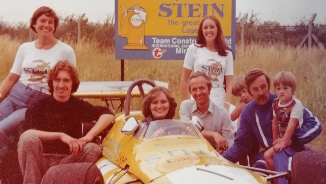 Group of six people, including children, gathered around a yellow race car with sponsorship decals on it, outdoors in a grassy field with a sign promoting Stein beer in the background. Chris Ward. Legacy Outlaws Racing Team.