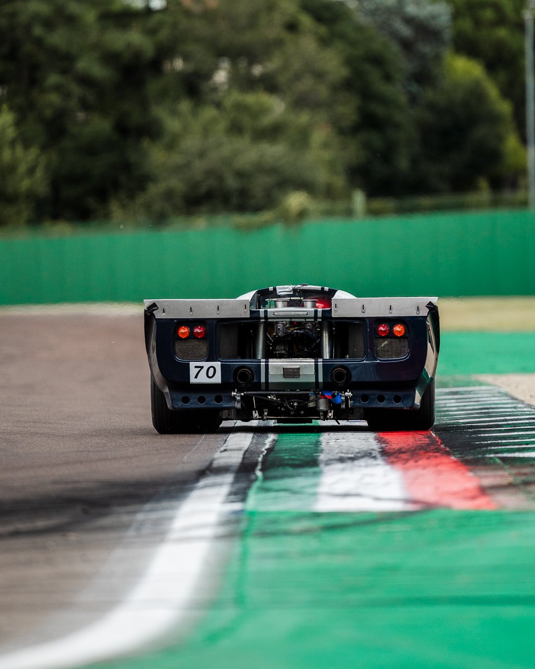 Rear view of a race car on a track, with green and red markings, and a green fence and trees in the background. Lola T70 MkIIIB Peter Auto Imola Classic 2025. Legacy Outlaws Racing Team.
