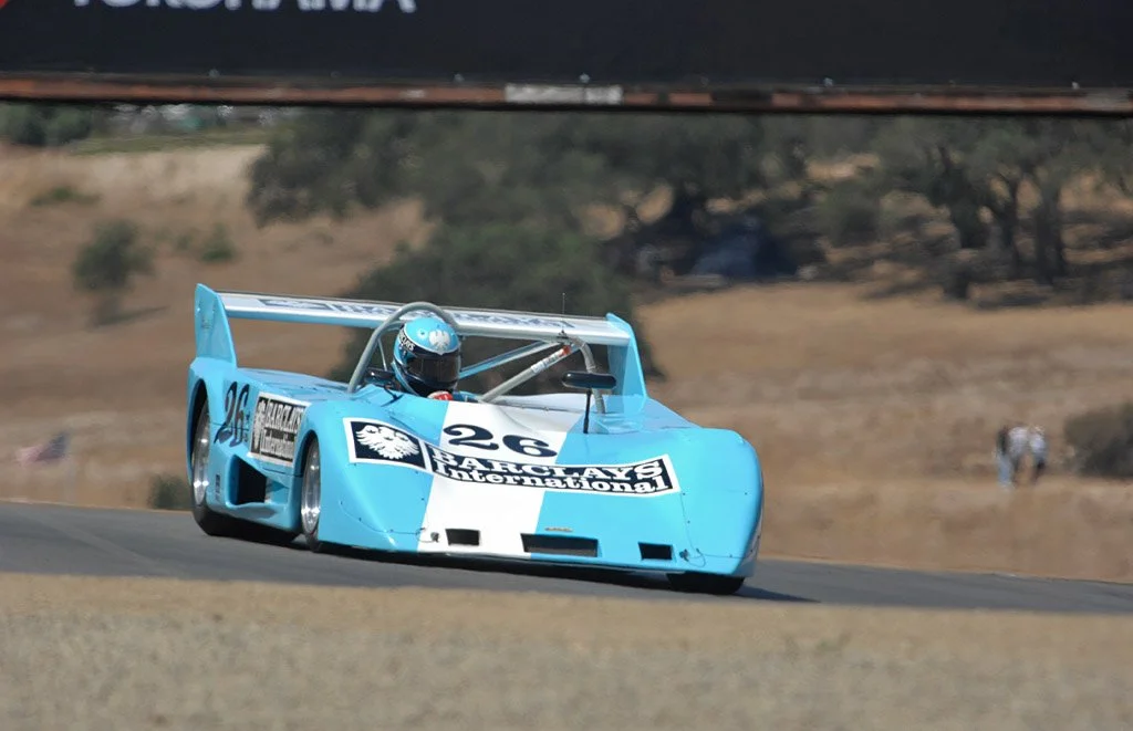 A blue vintage race car speeding on a track with a driver wearing a helmet. Lola T292 Peter Auto 2025. Legacy Outlaws Racing Team.