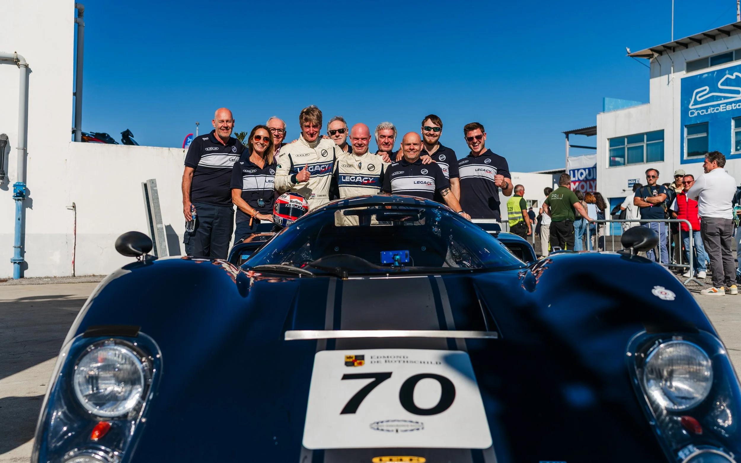 Group of race car drivers and team members in racing uniforms standing behind a navy Lola MkIIIb T70 race car with the number 70, on a pit lane at a motorsport event under clear blue skies in Estoril Cascais Peter Auto 2025