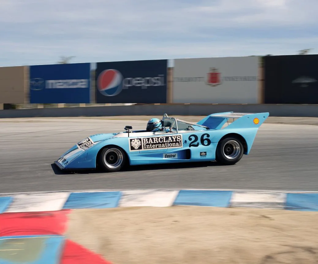 A vintage blue race car with the number 26 on its side speeding on a racetrack. Lola T292 Peter Auto 2025. Legacy Outlaws Racing Team.. The car has a large rear wing and is sponsored by Barclays and Castrol, with a Shell logo on the rear wing.