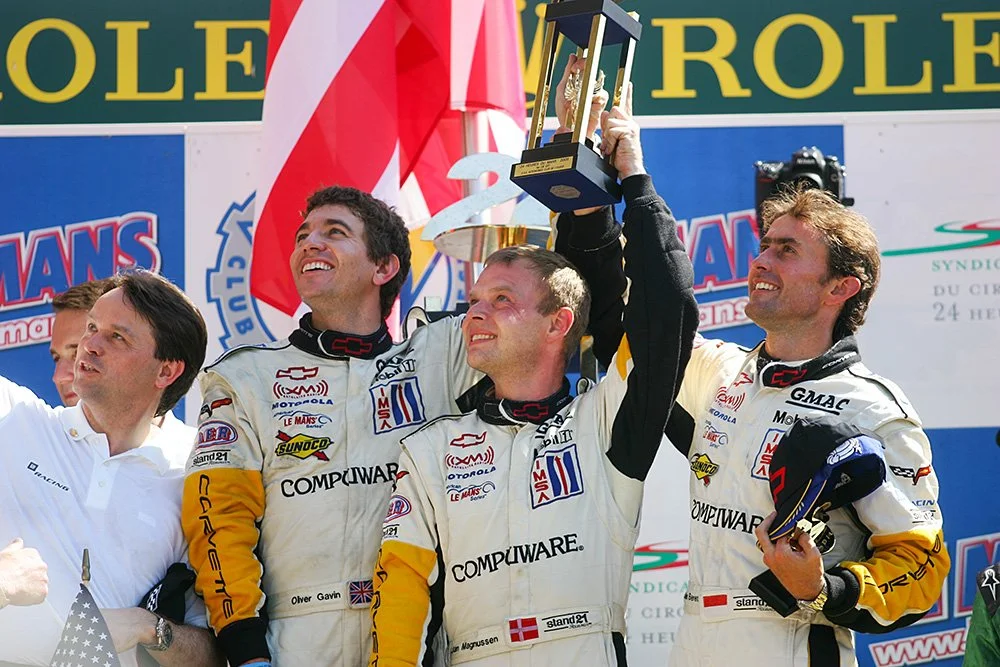 Four men celebrating a motorsports victory on the podium, with one holding a trophy above his head. They are wearing racing suits with various sponsor logos and standing in front of a backdrop with racing banners. Jan Magnussen. Le Mans 24hrs podium.