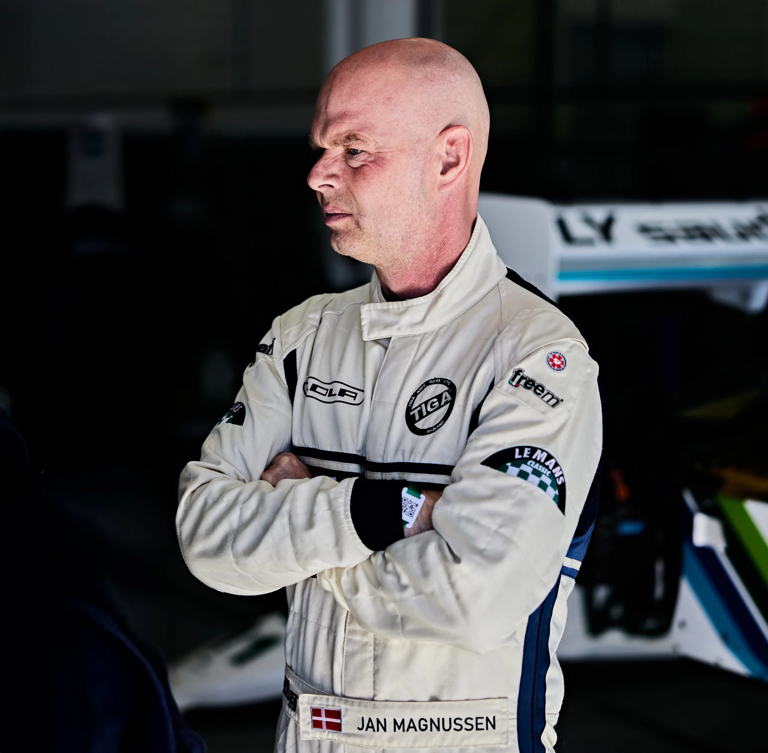 A male race car driver with a shaved head standing with arms crossed, wearing a white racing suit with various sponsor logos. Jan Magnussen. Legacy Outlaws Racing Team.