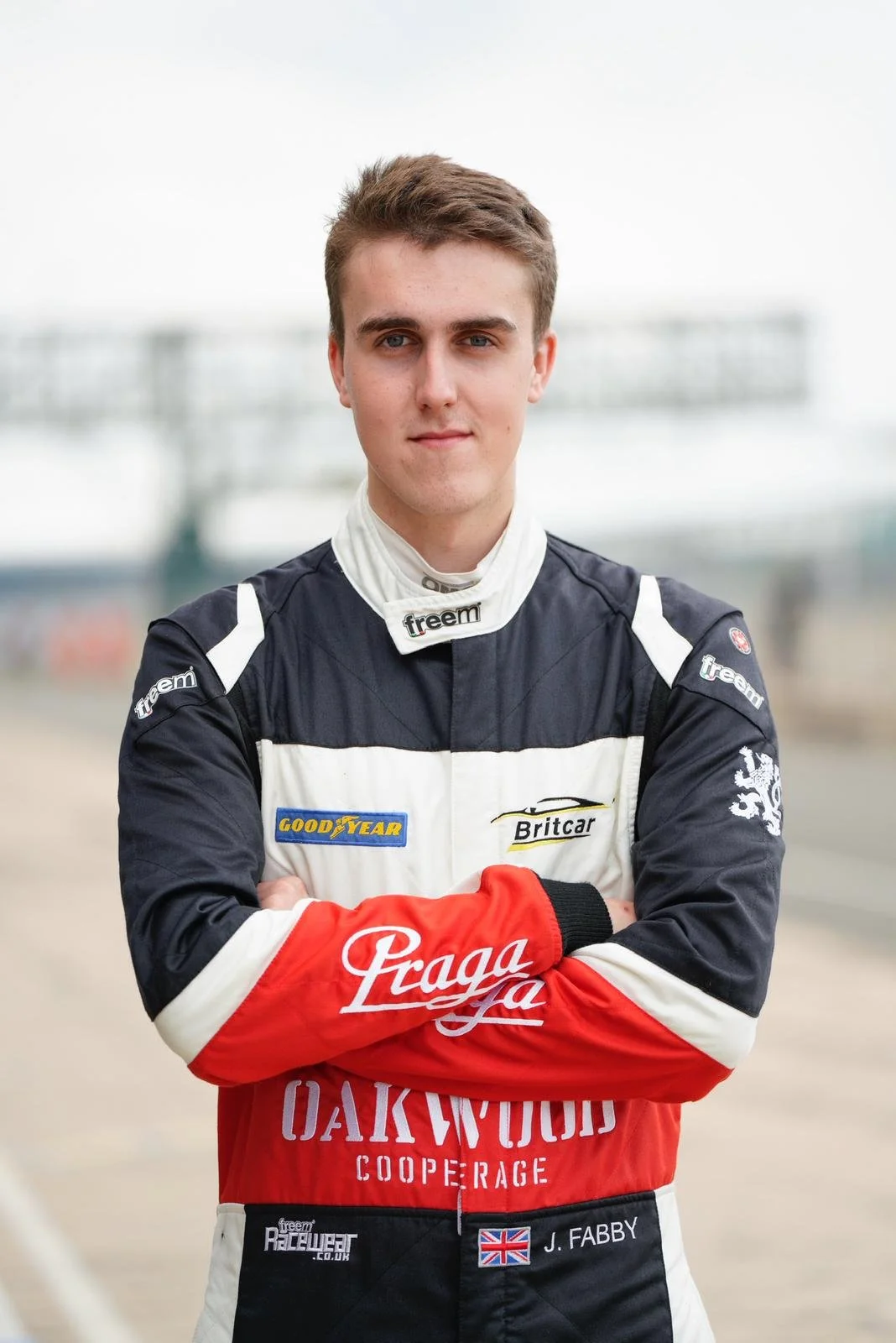 Young male race car driver in racing suit with arms crossed, standing outdoors on race track. Jack Fabby. Legacy Outlaws Racing Team. Prega.