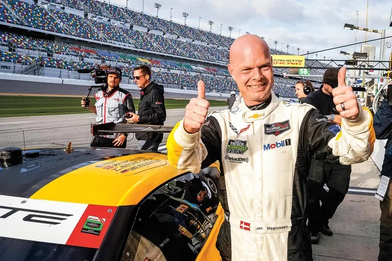 A race car driver in a white racing suit with sponsor logos smiles and gives two thumbs up at a racetrack, with other people and a grandstand in the background. Jan Magnussen. Daytona International Speedway. Daytona 24hrs. Legacy Outlaws Racing Team.