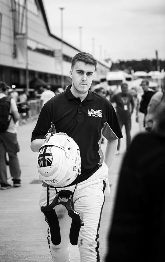 Young male race car driver holding a helmet with racing suit and in a busy pit lane or garage area at a racetrack. Jack Fabby. Legacy Outlaws Racing Team.