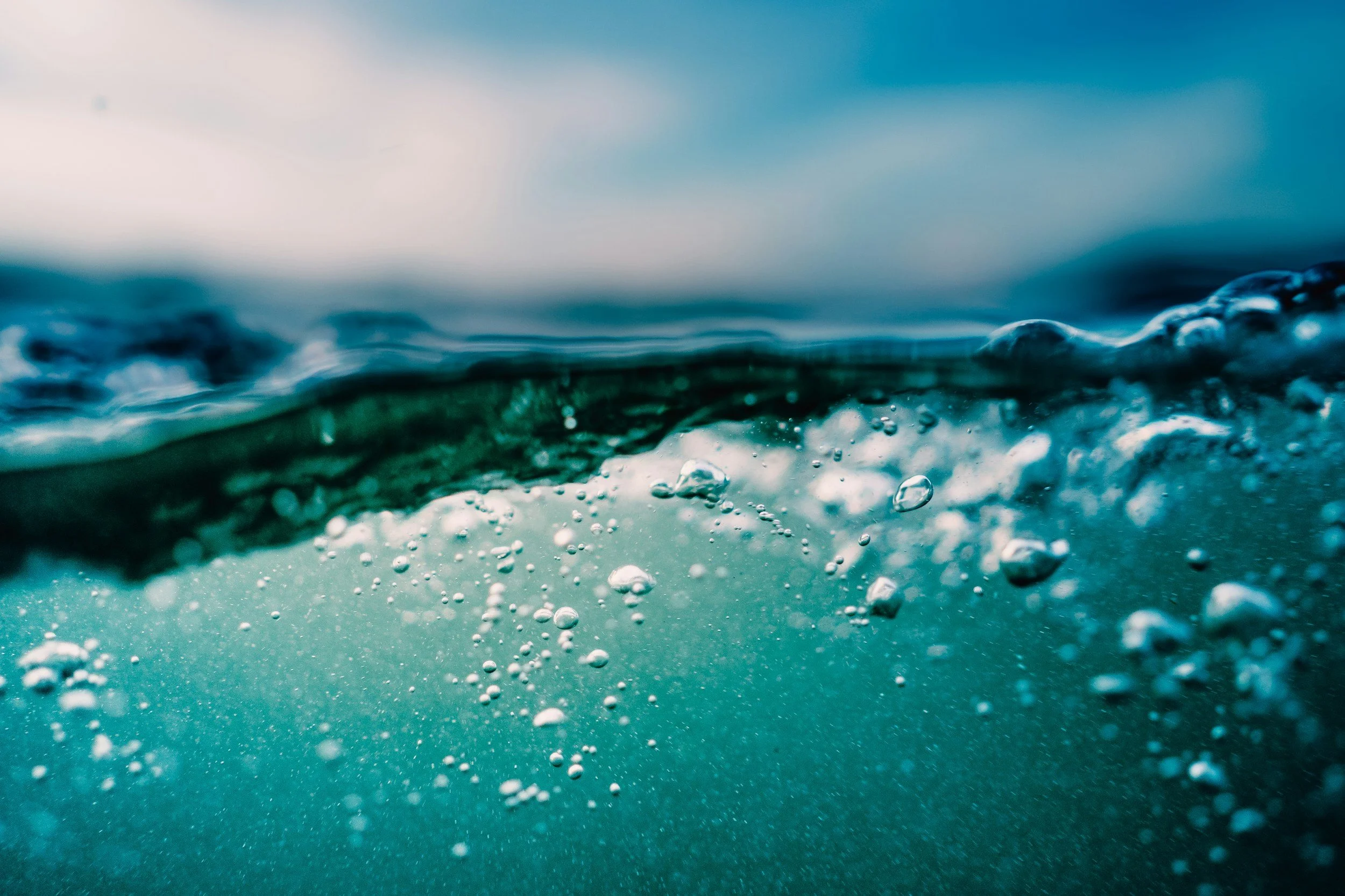 Close-up of ocean water with bubbles and waves, taken underwater with a blurred sky in the background.