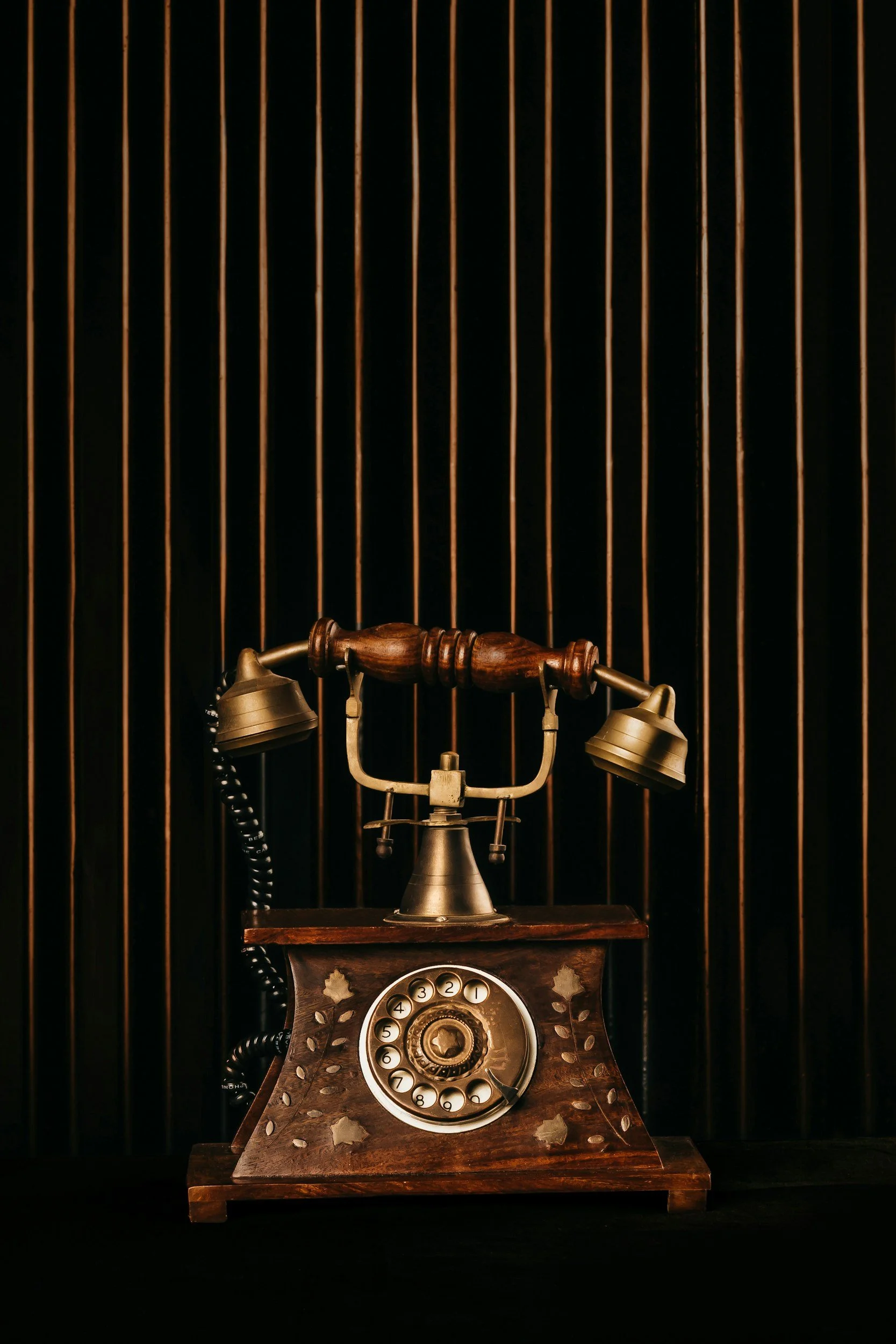 Vintage rotary telephone with brass and wood details placed on a dark surface, against a black background with gold vertical lines.