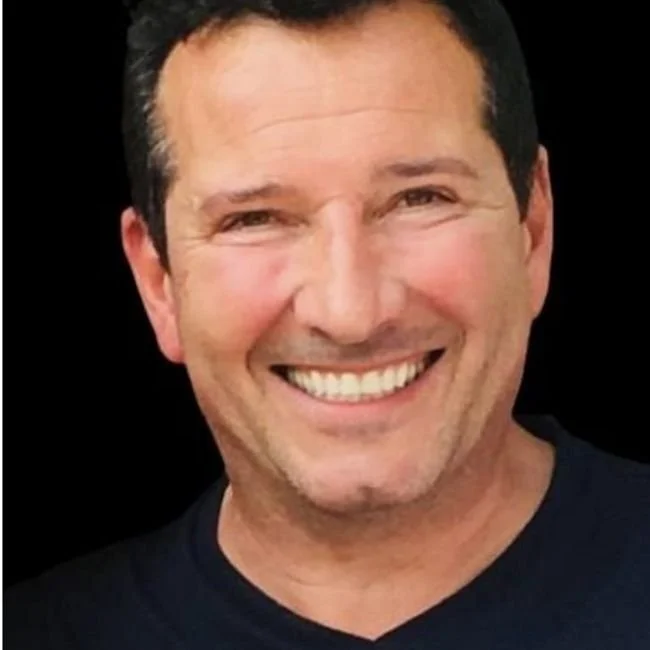 Close-up photo of a man smiling with short dark hair, wearing a black shirt, against a black background