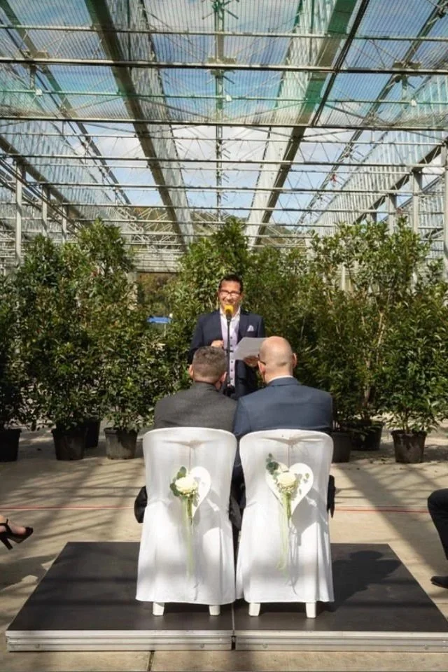 Eine Hochzeitzeremonie in einem Gewächshaus mit einem Mann, der vor einem Mikrofon und einer Lesung steht, während zwei Männer im Anzügen auf Stühlen sitzen. Die Stühle sind mit weißen Stoffen und Blumen dekoriert, im Hintergrund viele grüne Pflanzenund ein Glasdach, das Sonnenlicht durchlässt.