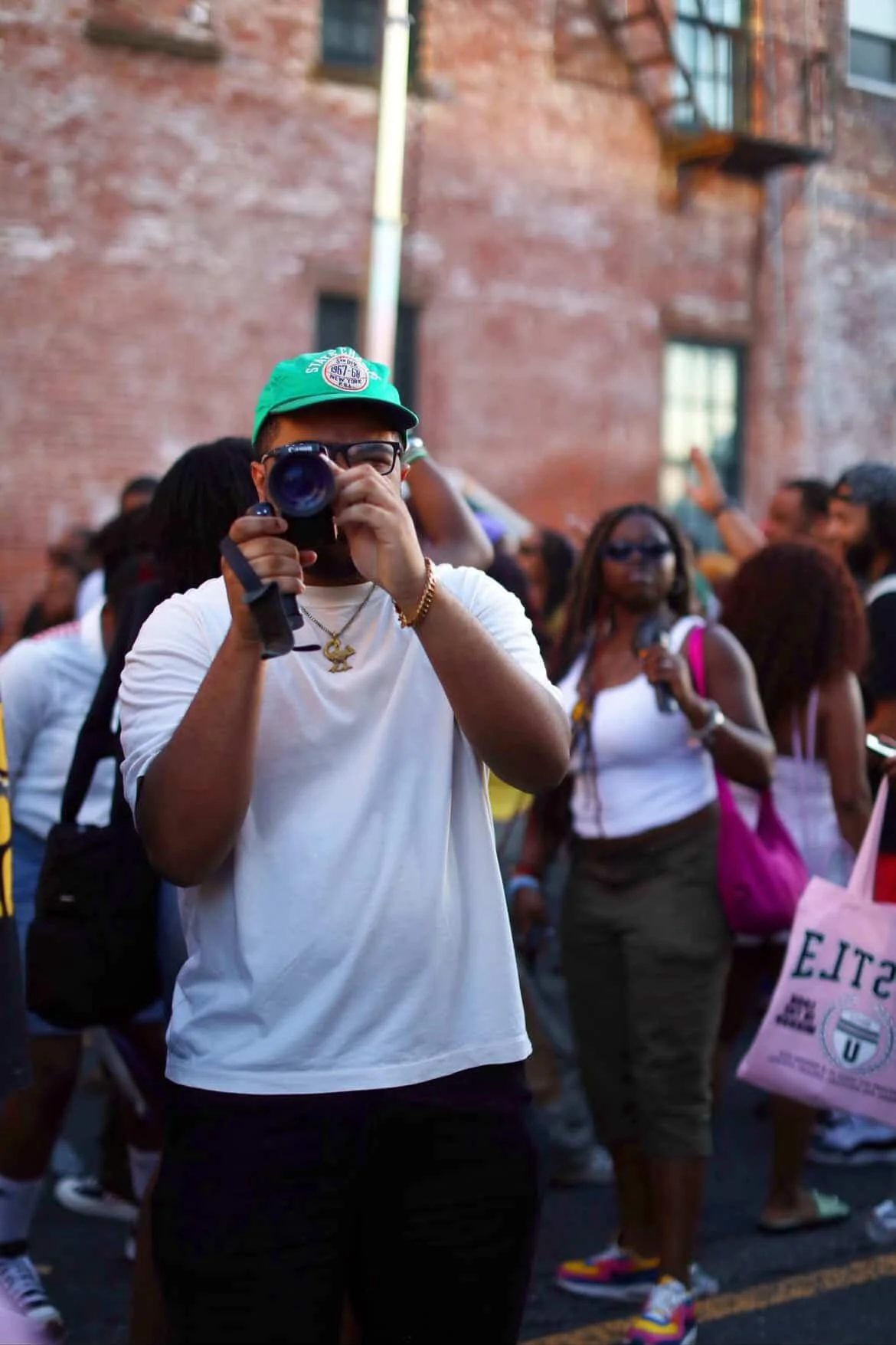 Person wearing glasses and a green cap takes a photo with a camera at a crowded outdoor event, with a brick building in the background.