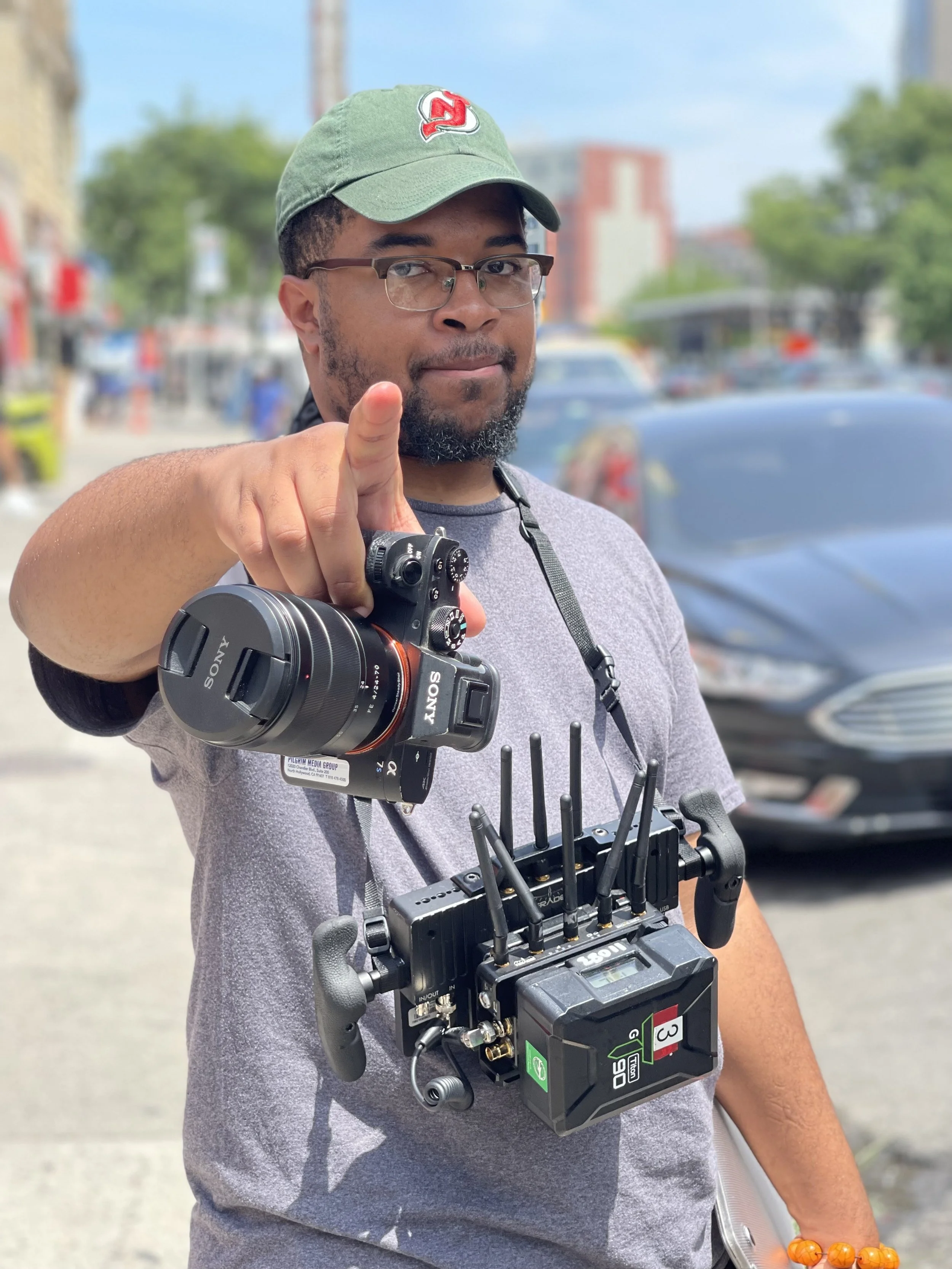A man with glasses and a baseball cap pointing towards the camera, holding a camera rig and a remote control, standing outdoors on a street with cars and buildings in the background.
