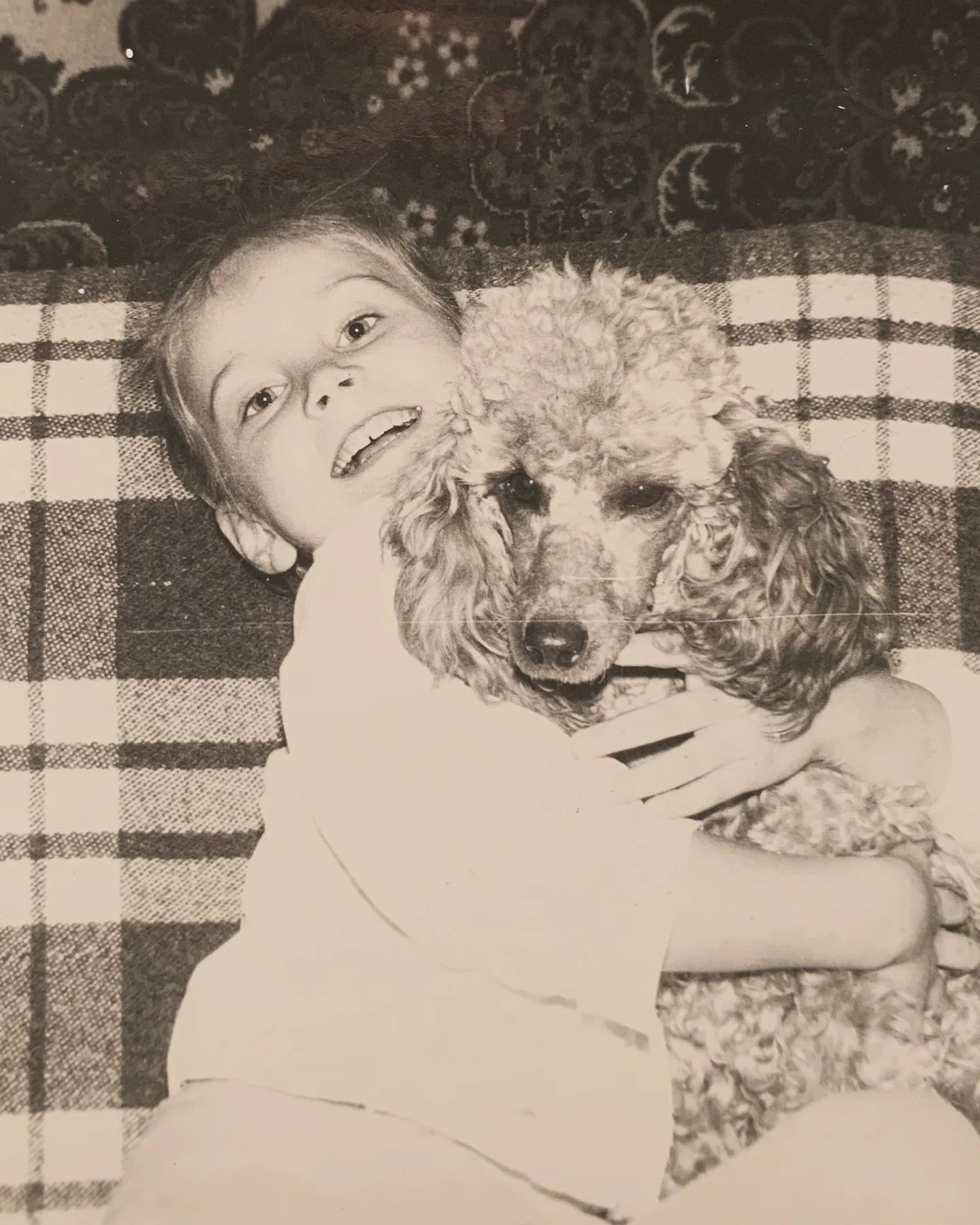 A young girl lying on her back hugging a poodle dog, both looking at the camera, on a patterned blanket.