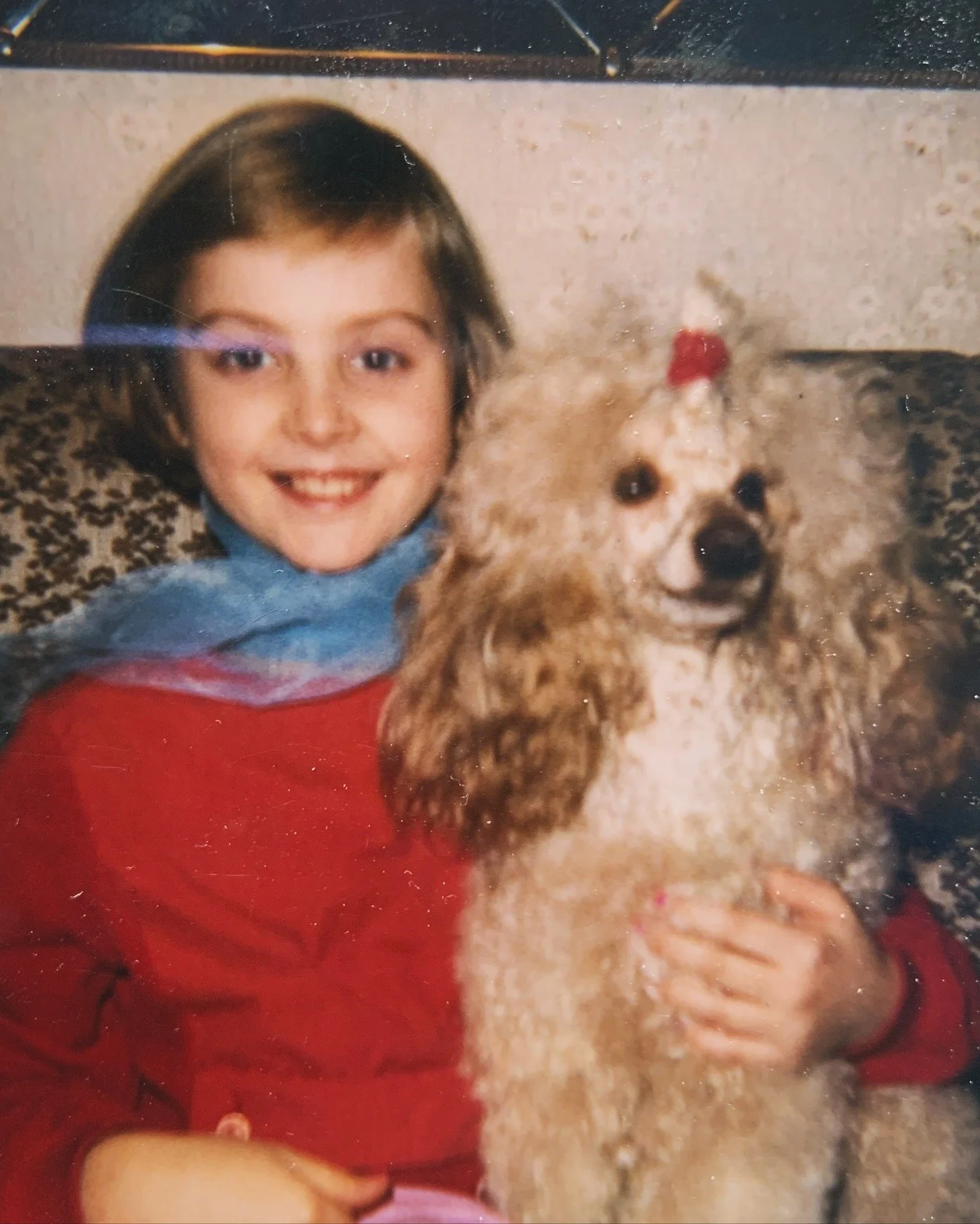 Child with short hair smiling and sitting next to a poodle with a red bow on its head, in a cozy indoor setting.