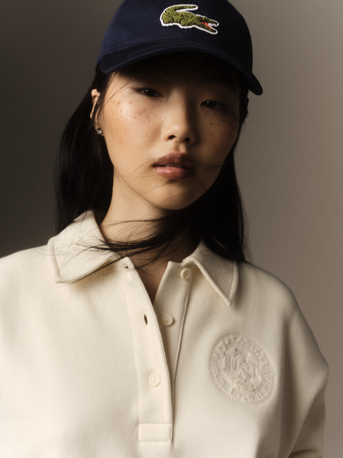 Jeune femme avec un chapeau bleu portant un logo de crocodile, portant un polo blanc avec un badge brodé, décorée de taches de rousseur.